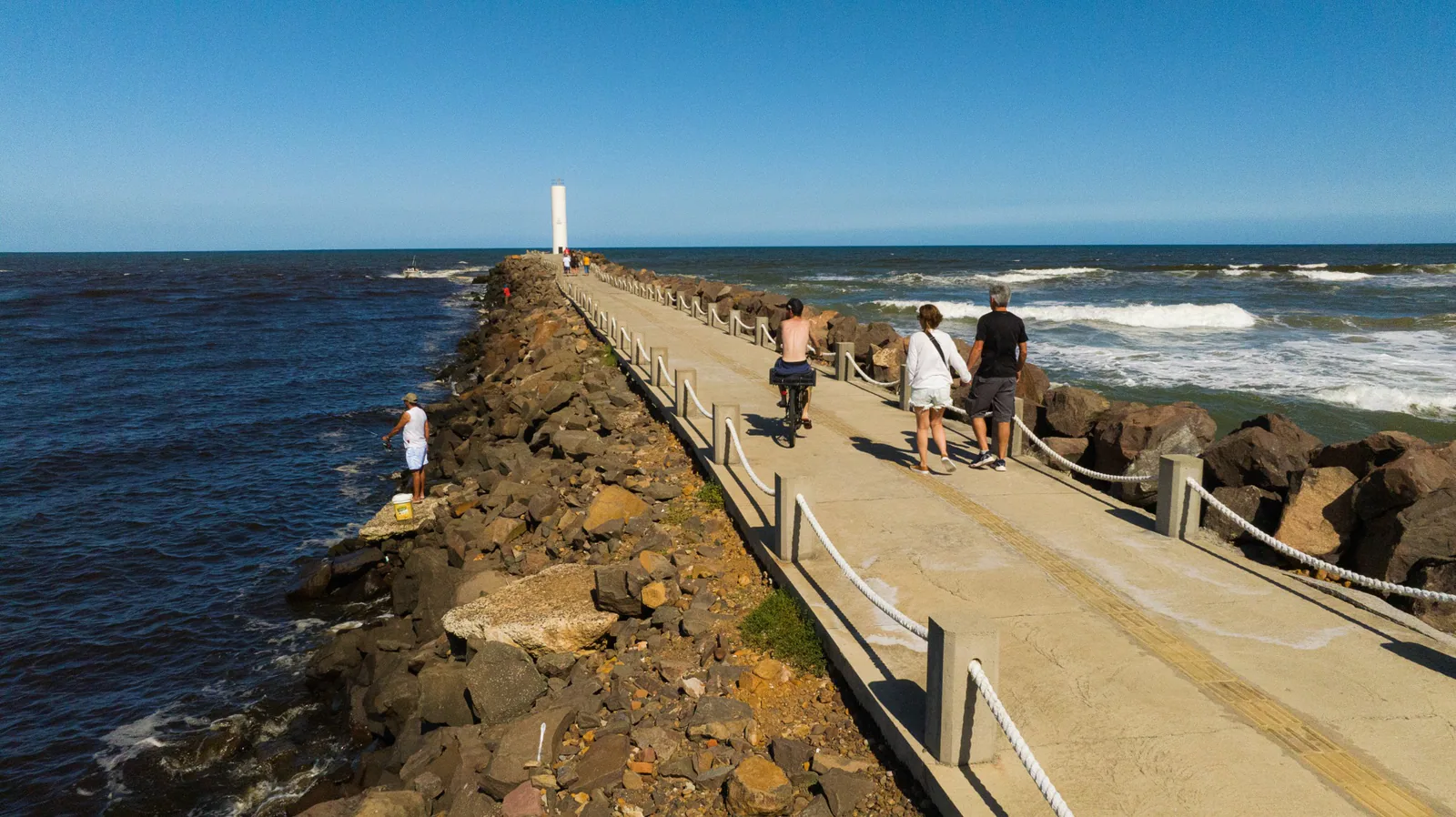 Personas caminando y en bicicleta por el espigón, con el faro y el mar al fondo