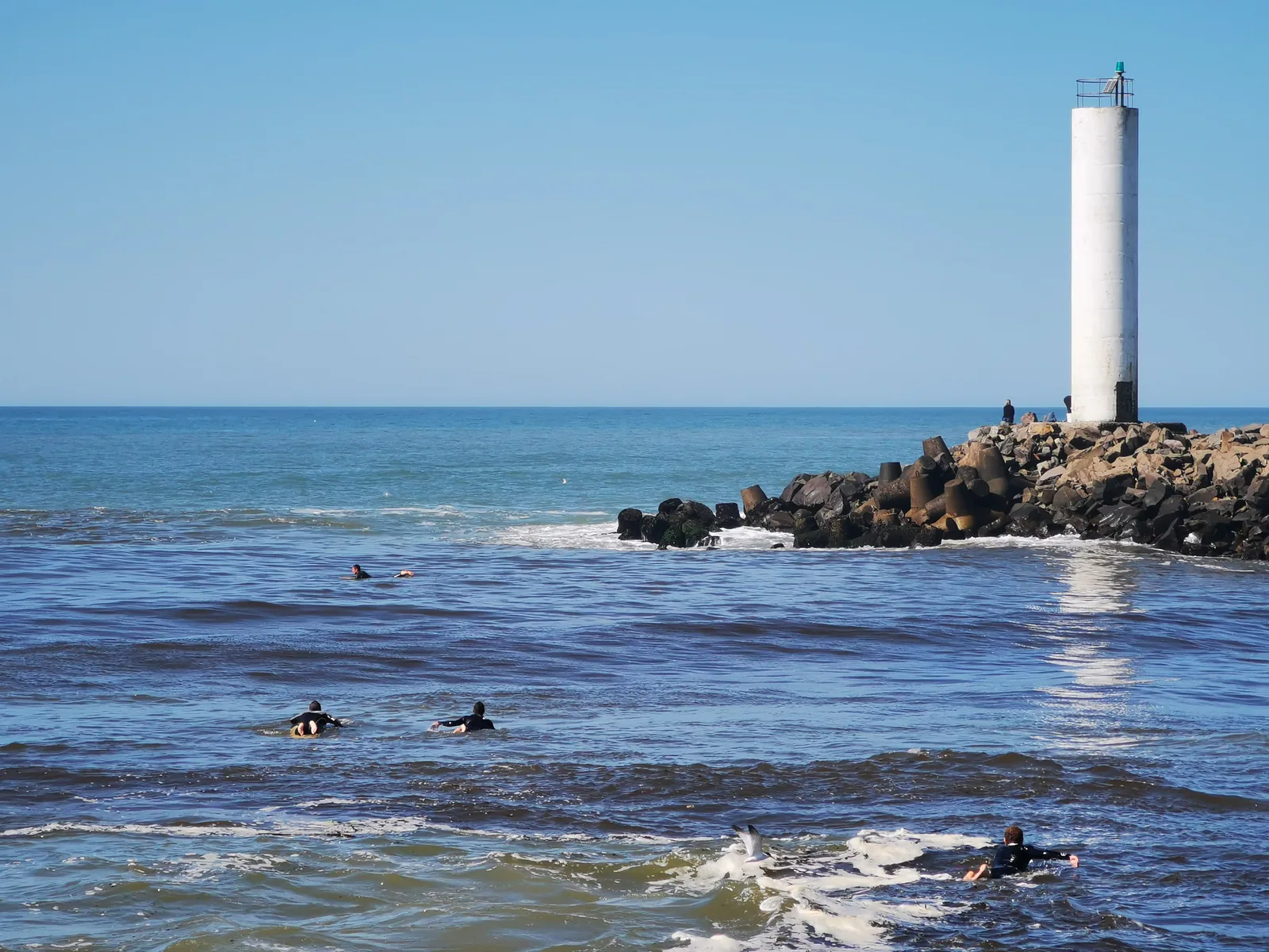 Surfistas en el mar junto al faro y las rocas de los Molhes da Barra