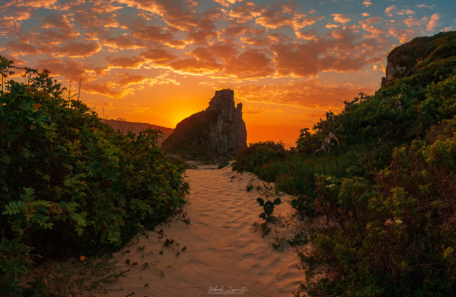 Caminho de areia entre a vegetação e o Morro da Guarita, sob céu dourado
