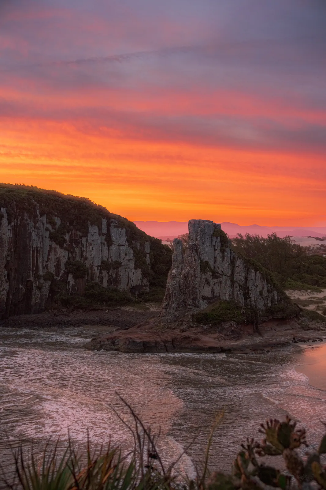 Morro da Guarita junto ao mar, com paredão rochoso ao lado e céu em tons de rosa e laranja